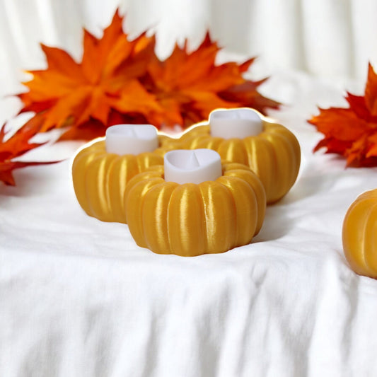 Three pumpkin-shaped candle holders with white candles on a white surface with orange leaves in the background.