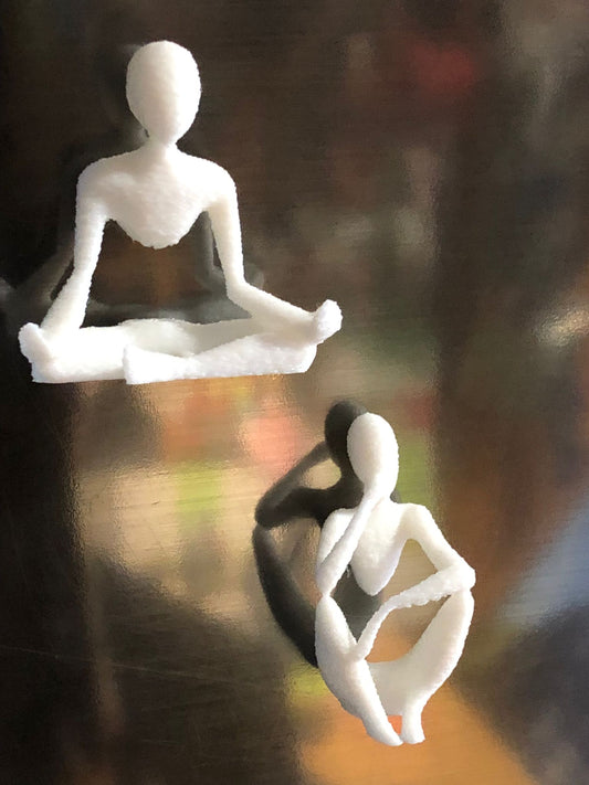 Set of two white 3D printed fridge magnets shaped like minimalist faceless human figures in yoga and meditation poses, displayed on a metallic surface.