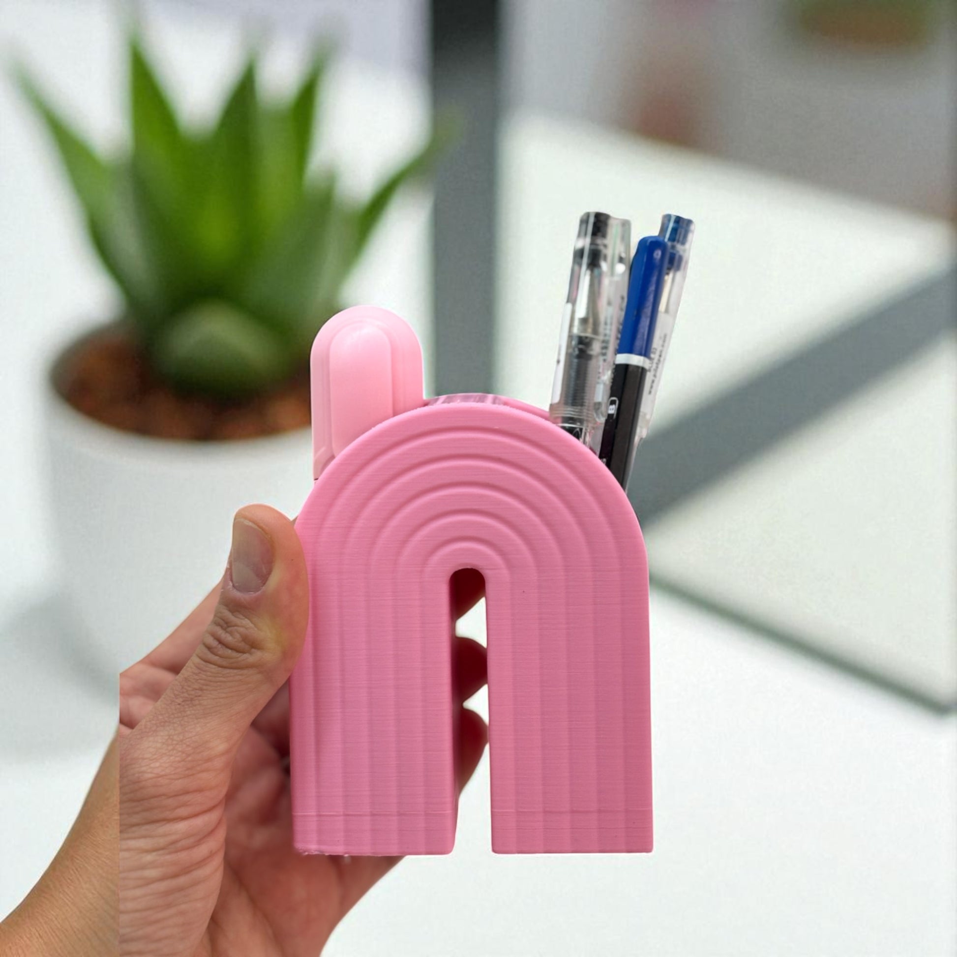Colorful 3D printed rainbow-shaped organizer used as a bookend, pen holder, and mini vase, displayed in two sizes on a shelf with books, stationery, and dried flowers.