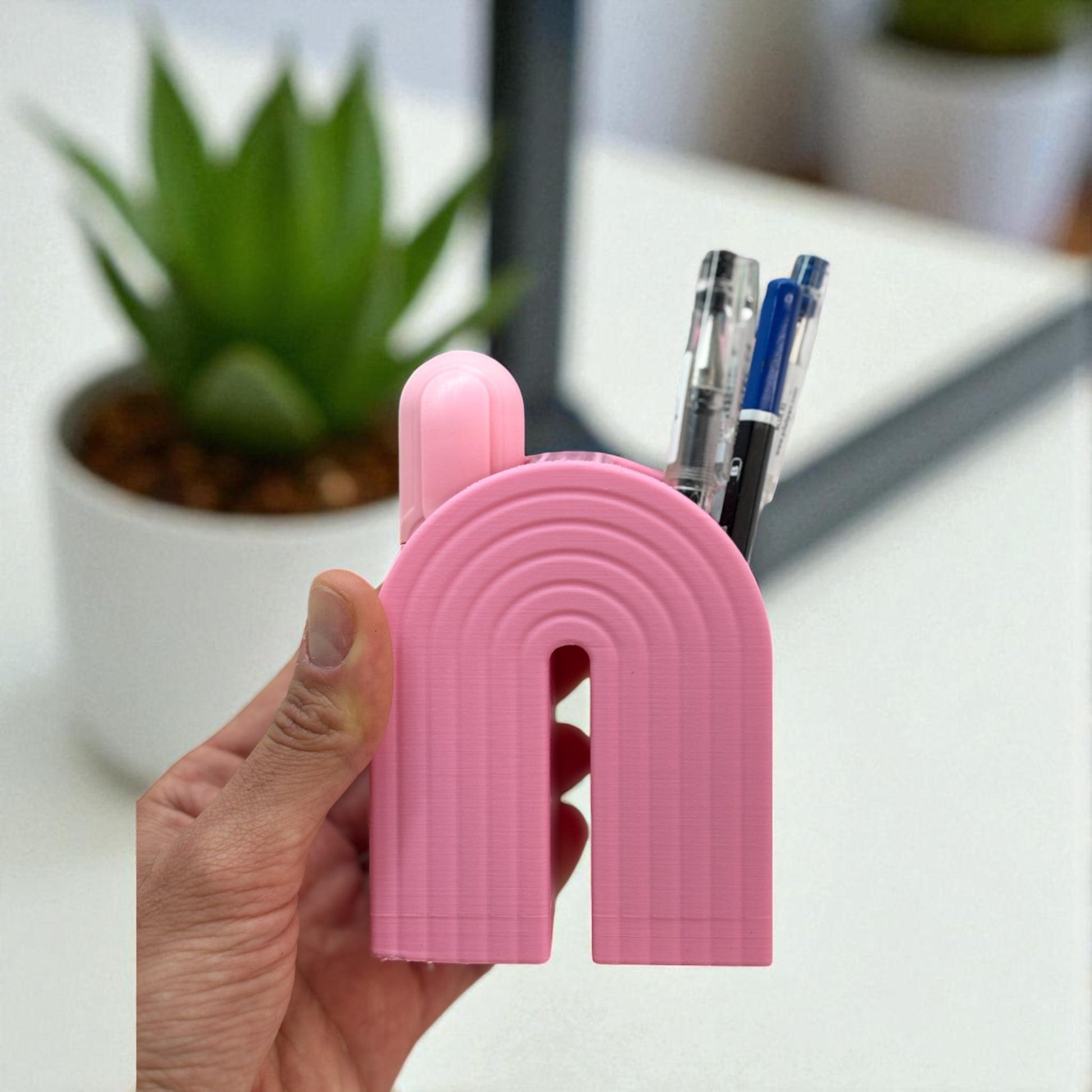 Colorful 3D printed rainbow-shaped organizer used as a bookend, pen holder, and mini vase, displayed in two sizes on a shelf with books, stationery, and dried flowers.
