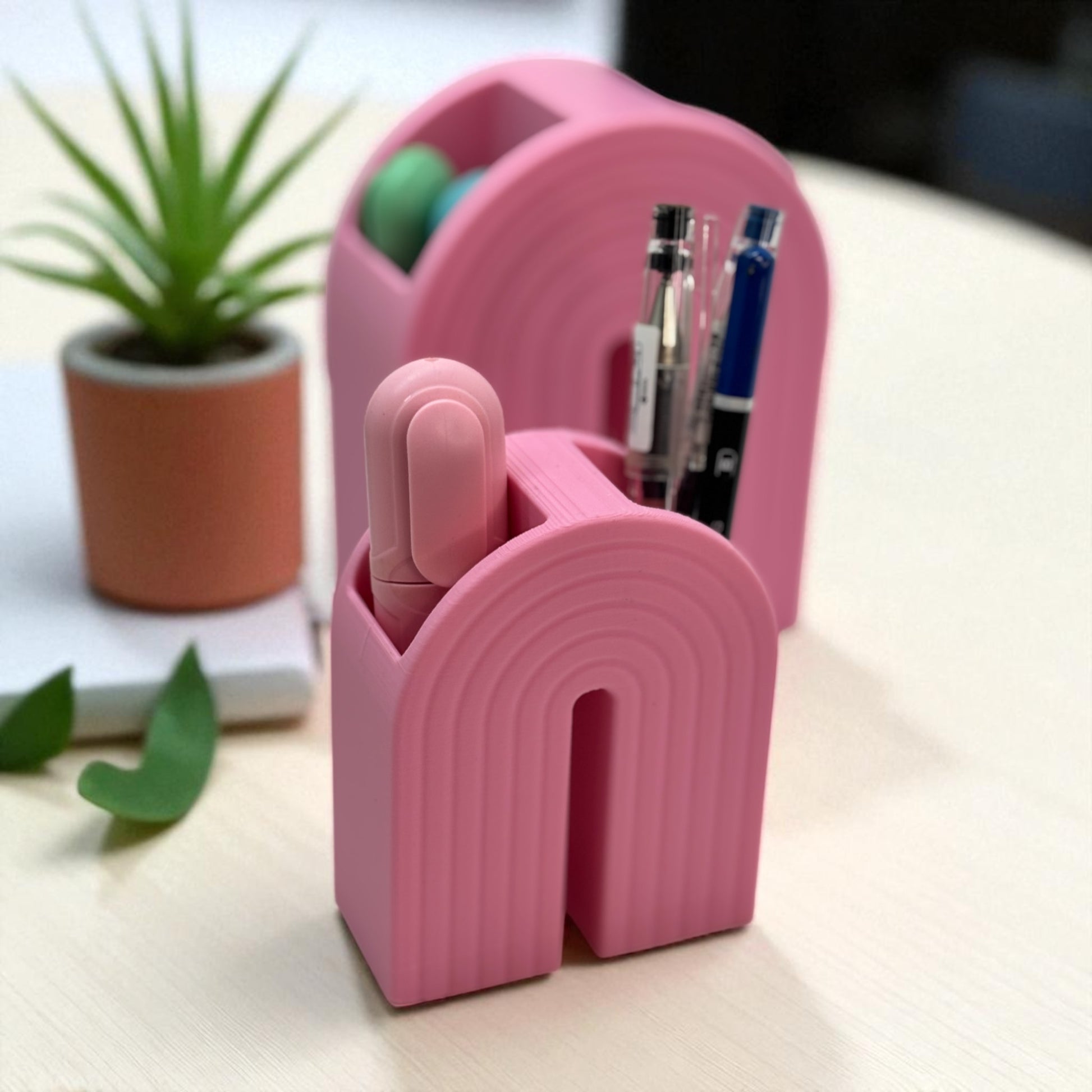 Colorful 3D printed rainbow-shaped organizer used as a bookend, pen holder, and mini vase, displayed in two sizes on a shelf with books, stationery, and dried flowers.