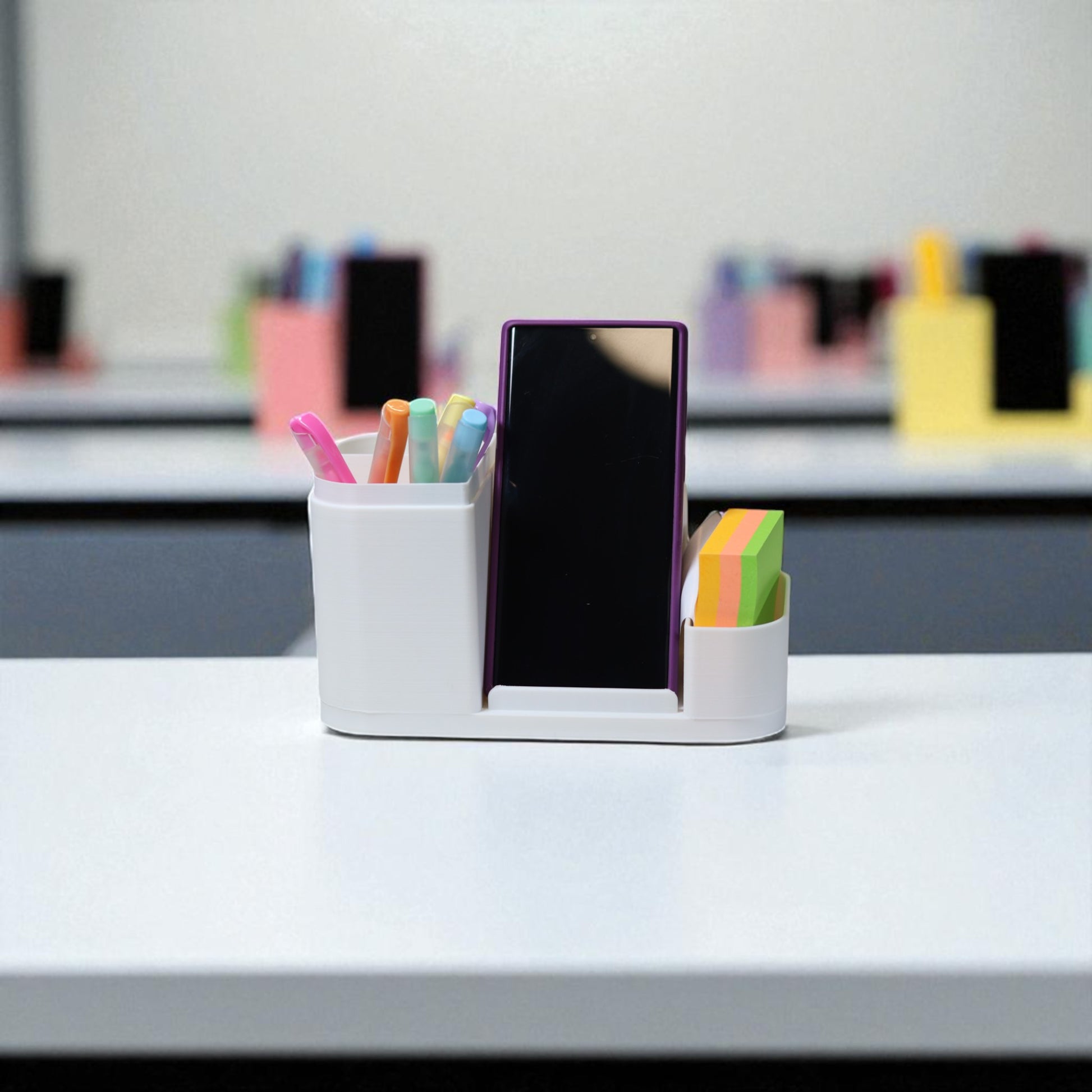 Kids desk organiser with phone stand, pencil/brush holder and sticky-note slot, 20×11×10 cm—white.Front view: organiser holding markers, smartphone in stand, and 3×3 sticky notes—back-to-school desk tidy.