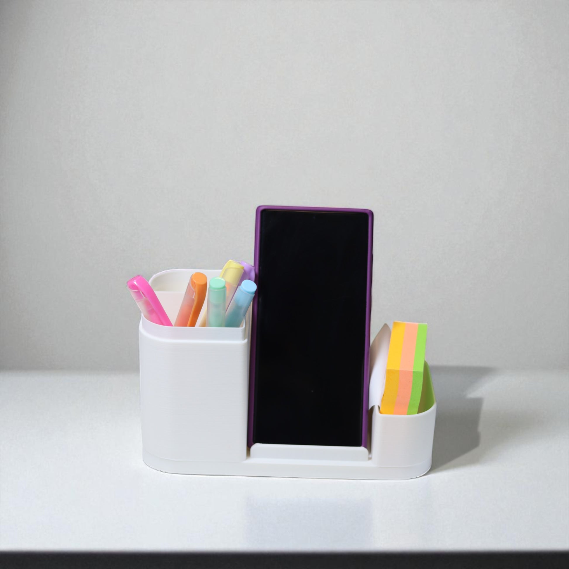 Kids desk organiser with phone stand, pencil/brush holder and sticky-note slot, 20×11×10 cm—white.Front view: organiser holding markers, smartphone in stand, and 3×3 sticky notes—back-to-school desk tidy.