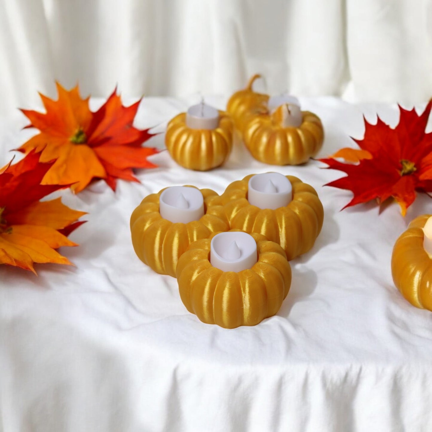 set of three pumpkin tealight holders (16×14×5.5 cm), neutral colours, showing option with led tealights lit, displayed on fall tabletop with autumn leaves.