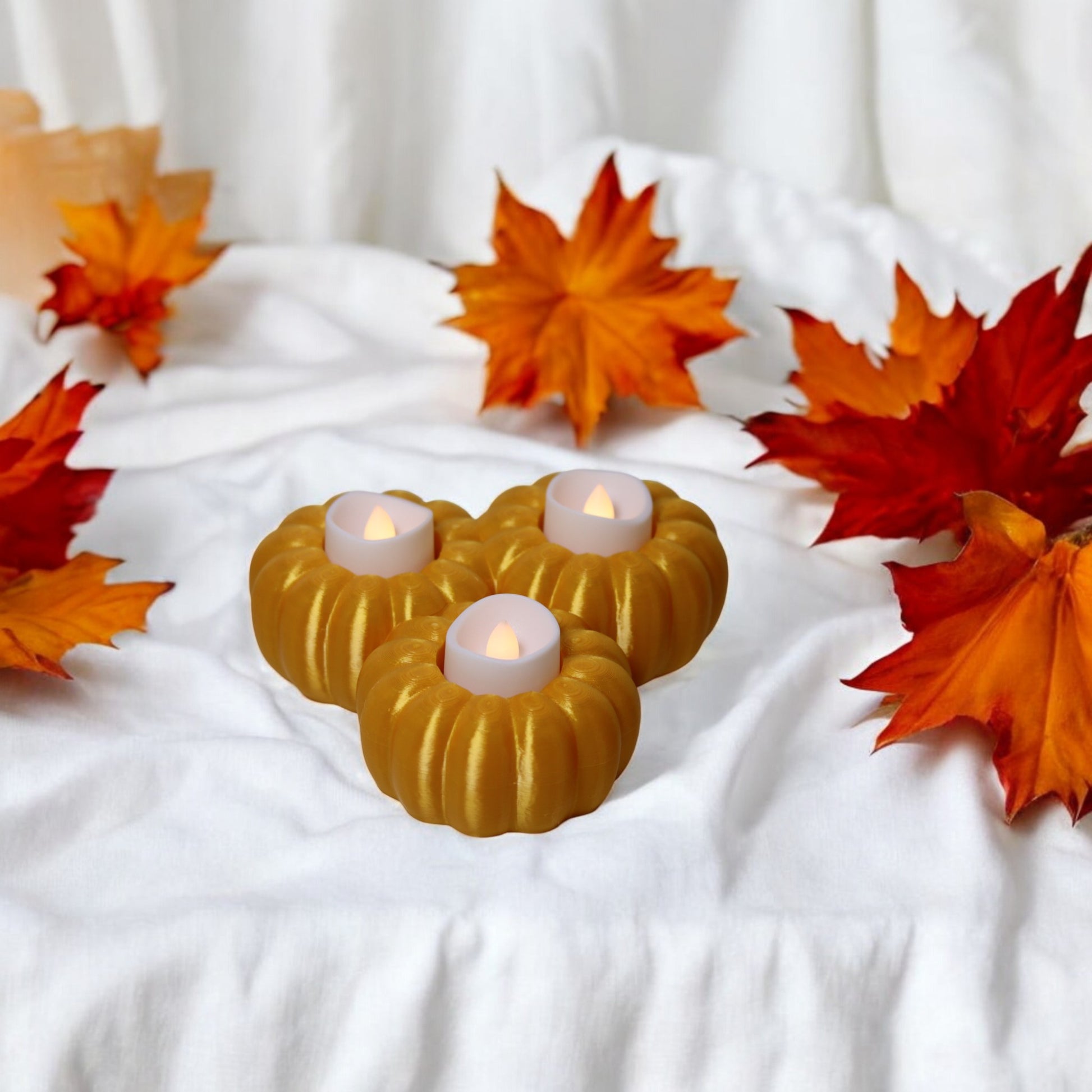 Set of three pumpkin tealight holders (16×14×5.5 cm), neutral colours, showing option with LED tealights lit, displayed on fall tabletop with autumn leaves.