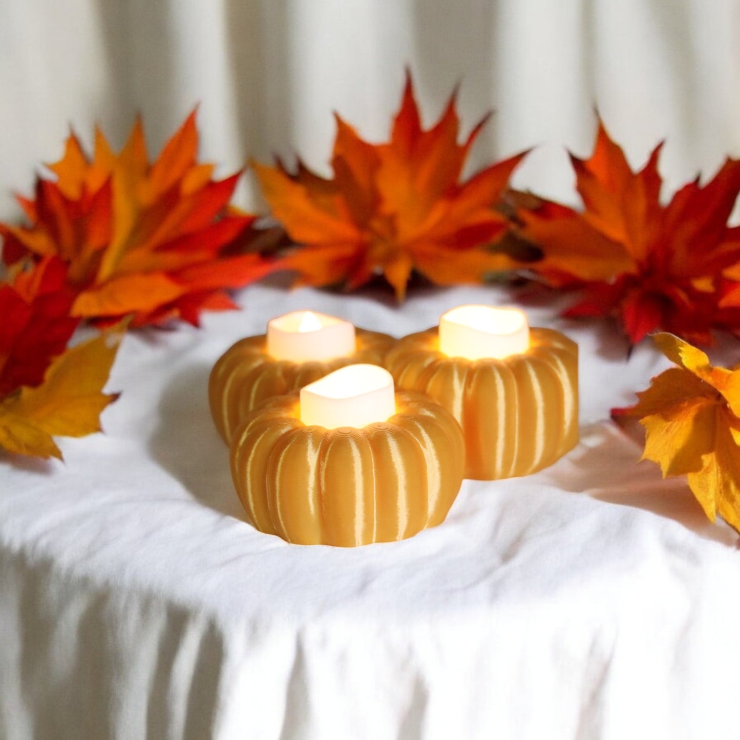 set of three pumpkin tealight holders (16×14×5.5 cm), neutral colours, showing option with led tealights lit, displayed on fall tabletop with autumn leaves.