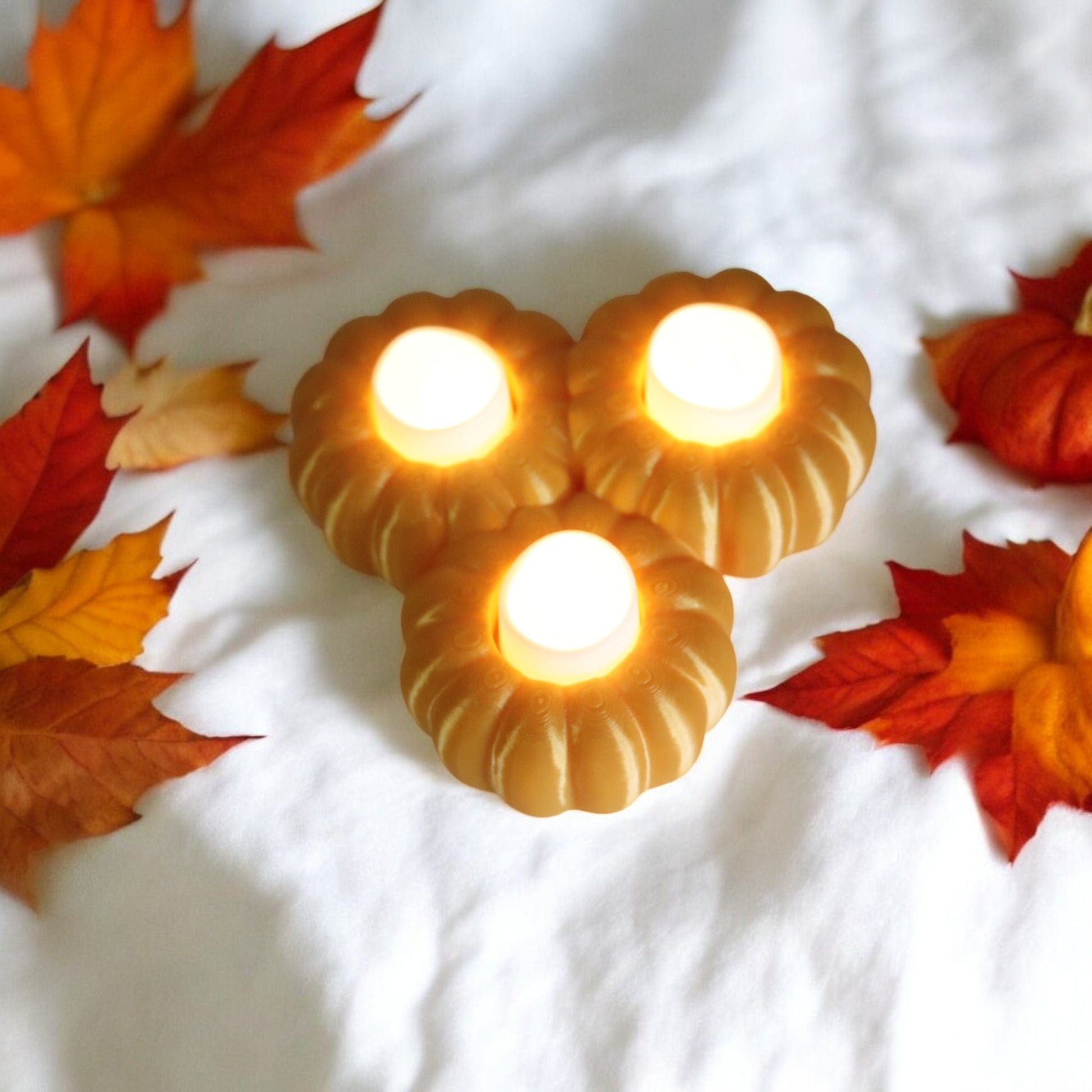 Set of three pumpkin tealight holders (16×14×5.5 cm), neutral colours, showing option with LED tealights lit, displayed on fall tabletop with autumn leaves.