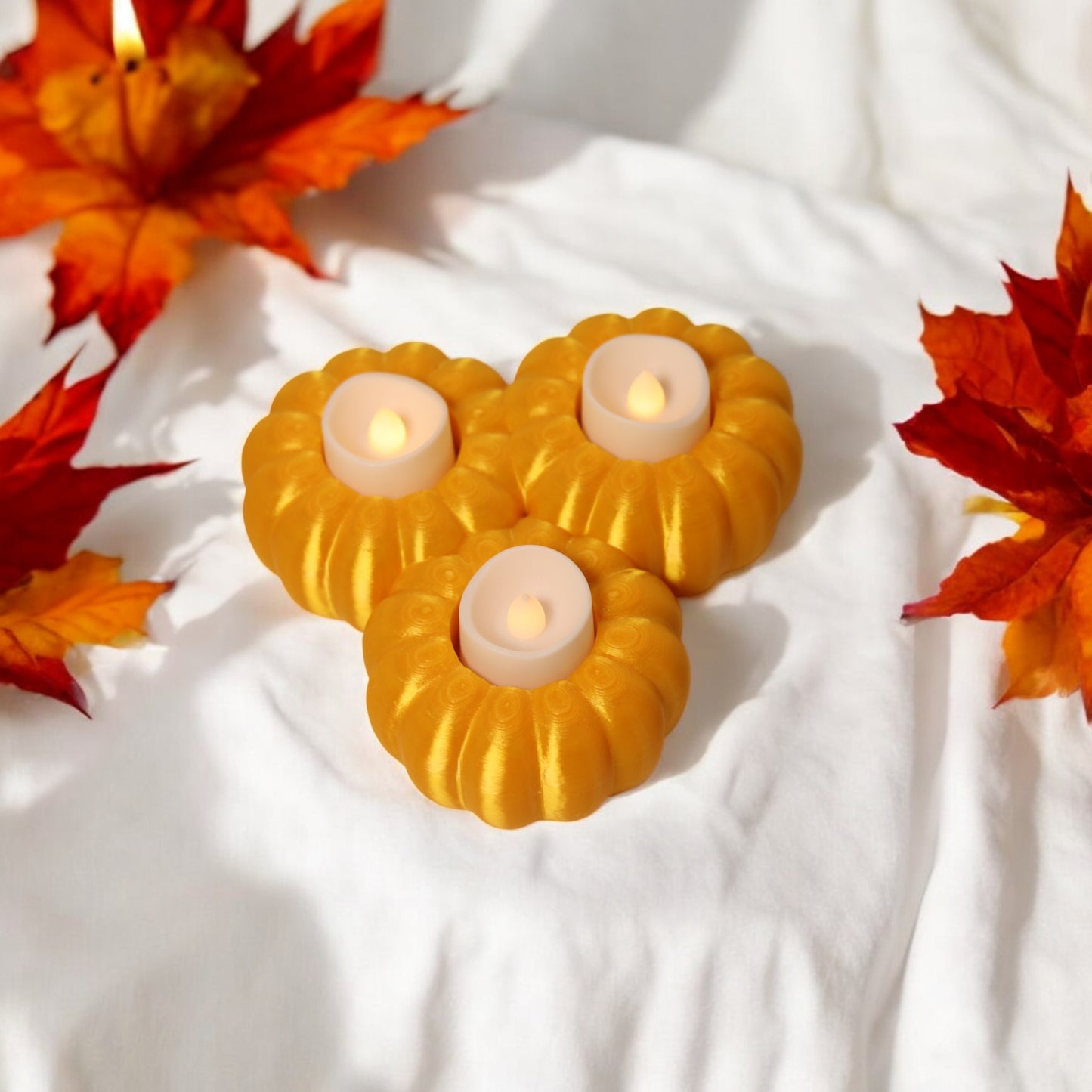 set of three pumpkin tealight holders (16×14×5.5 cm), neutral colours, showing option with led tealights lit, displayed on fall tabletop with autumn leaves.