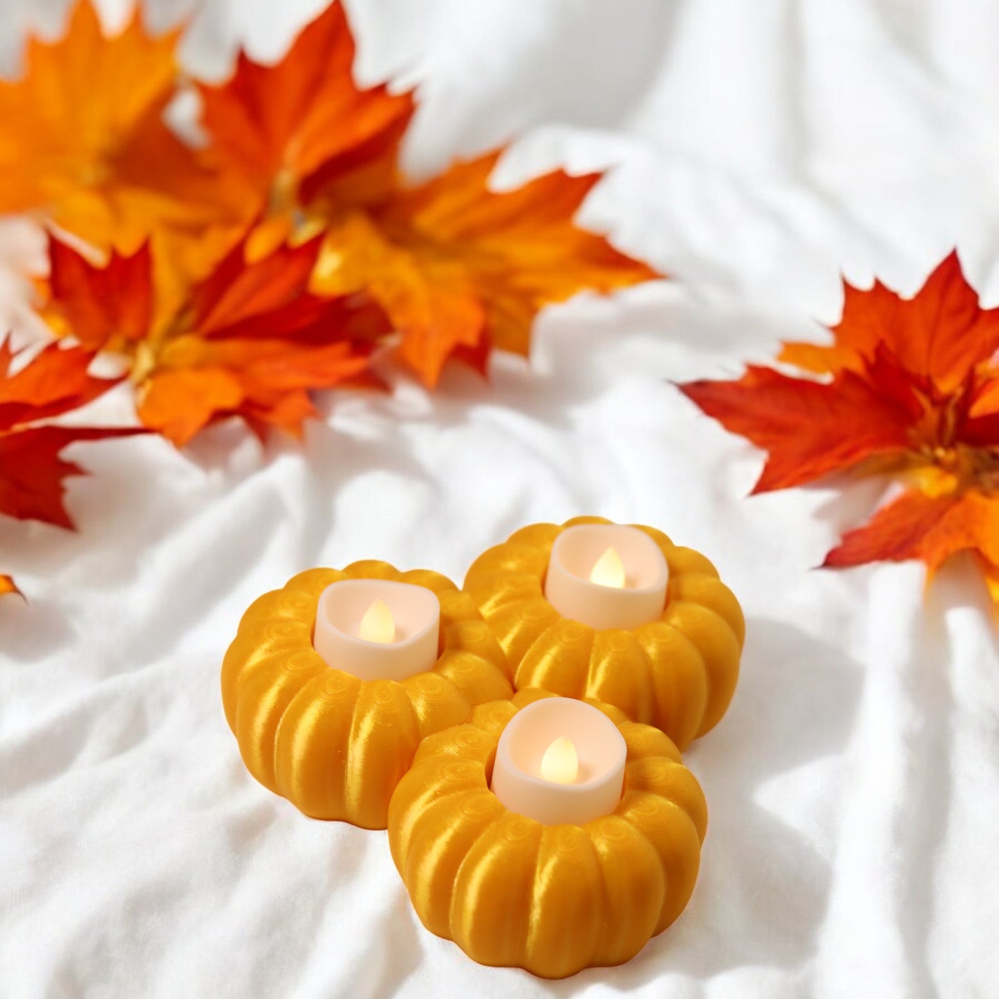 set of three pumpkin tealight holders (16×14×5.5 cm), neutral colours, showing option with led tealights lit, displayed on fall tabletop with autumn leaves.
