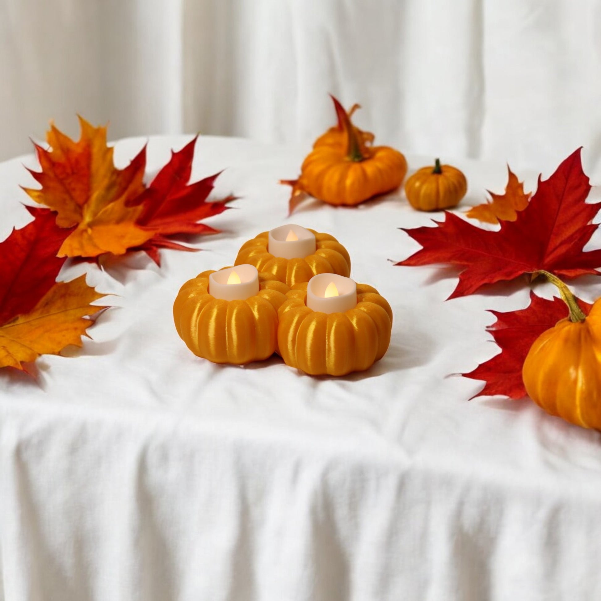 Set of three pumpkin tealight holders (16×14×5.5 cm), neutral colours, showing option with LED tealights lit, displayed on fall tabletop with autumn leaves.