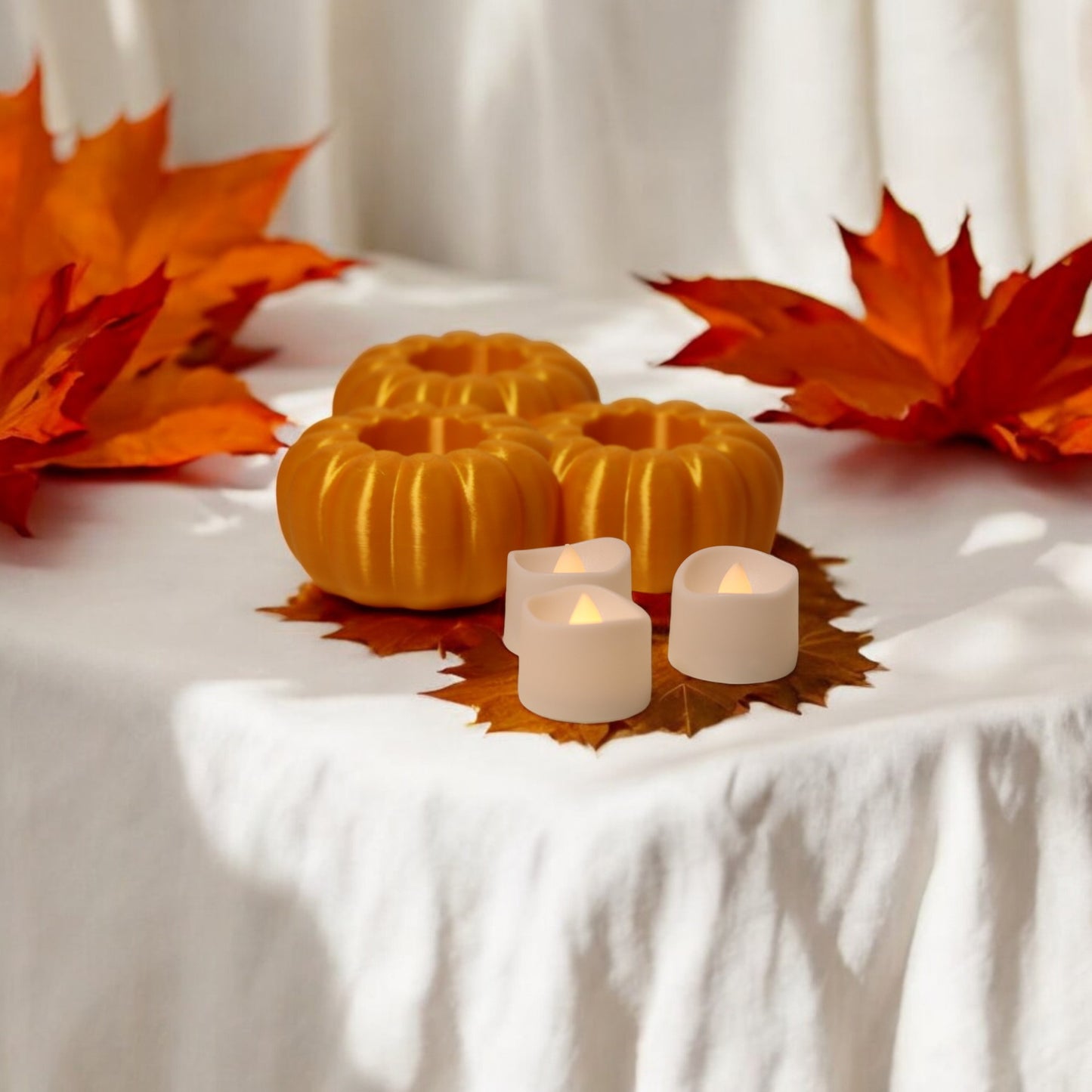 set of three pumpkin tealight holders (16×14×5.5 cm), neutral colours, showing option with led tealights lit, displayed on fall tabletop with autumn leaves.