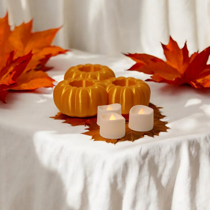 Set of three pumpkin tealight holders (16×14×5.5 cm), neutral colours, showing option with LED tealights lit, displayed on fall tabletop with autumn leaves.
