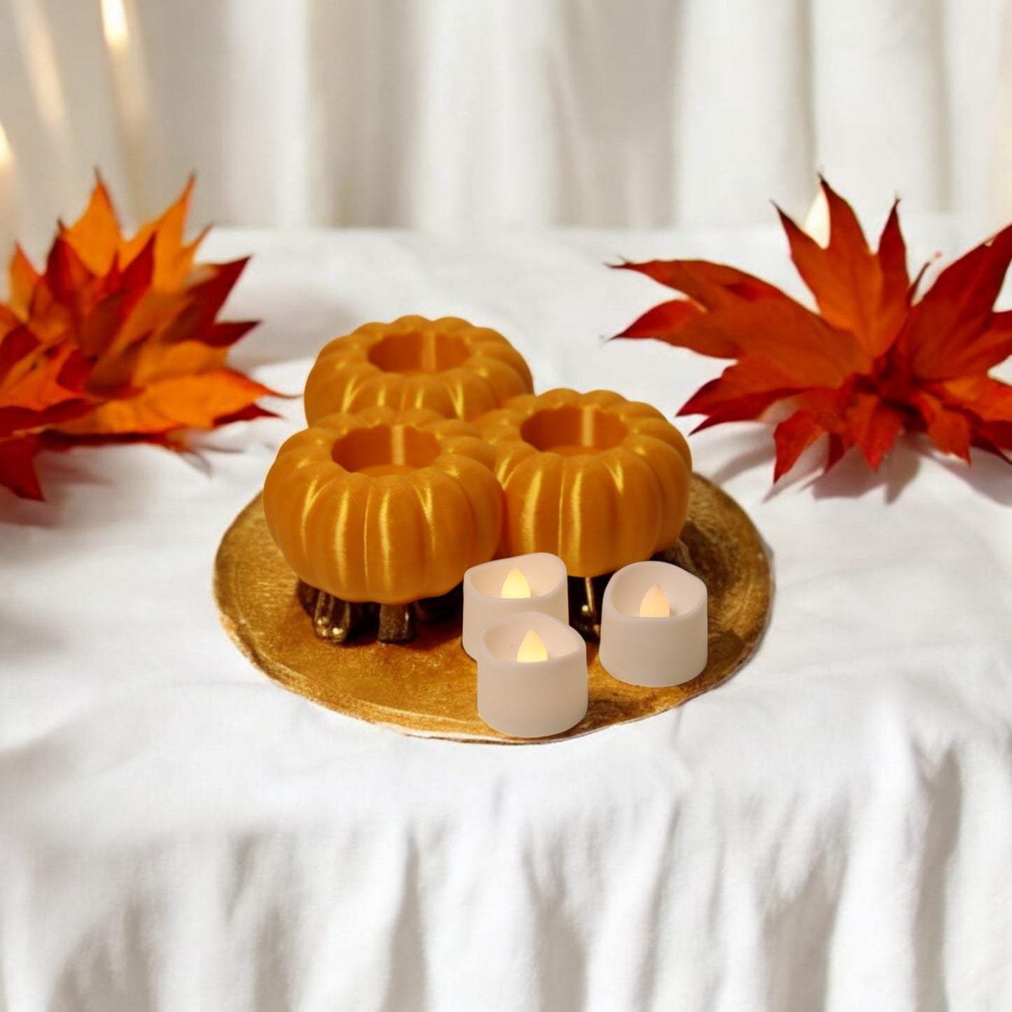 set of three pumpkin tealight holders (16×14×5.5 cm), neutral colours, showing option with led tealights lit, displayed on fall tabletop with autumn leaves.