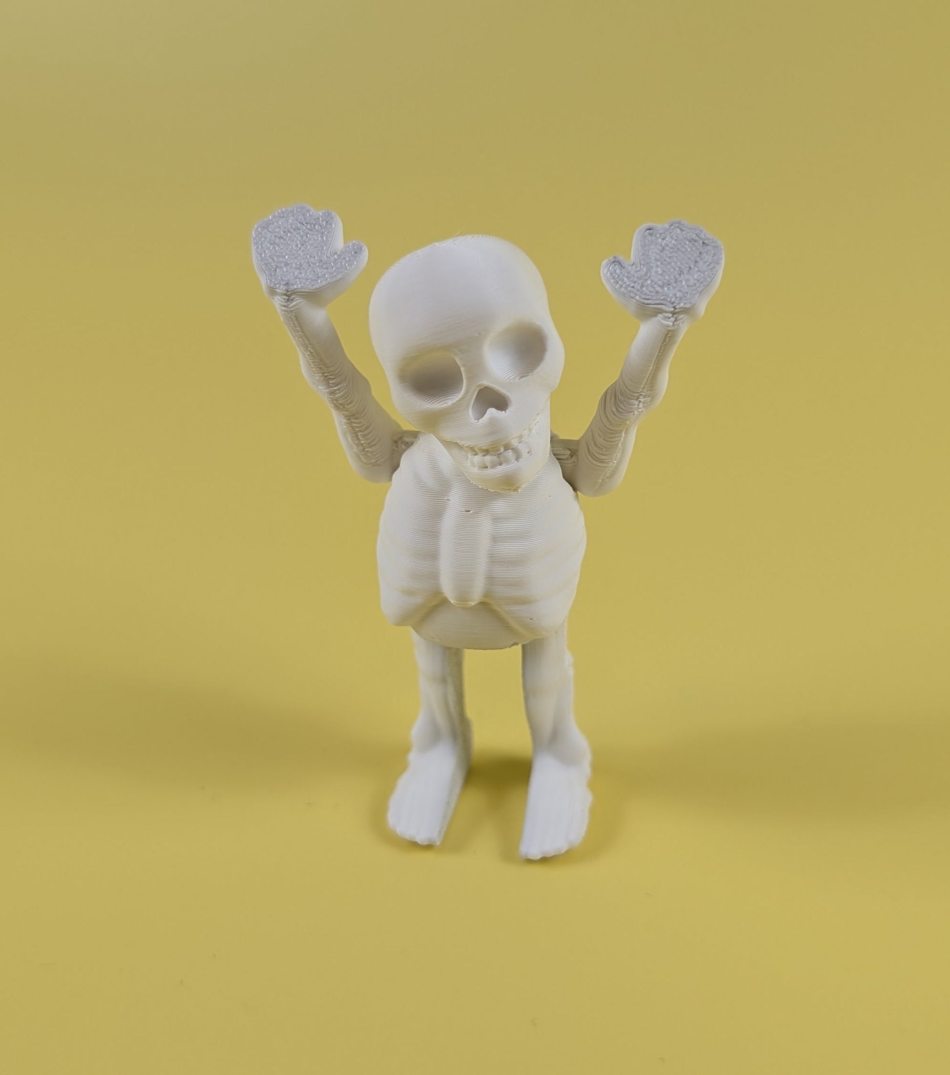 White 3D-printed skeleton figurine raising both arms on a yellow background; spooky-cute desk buddy with lightly poseable joints.