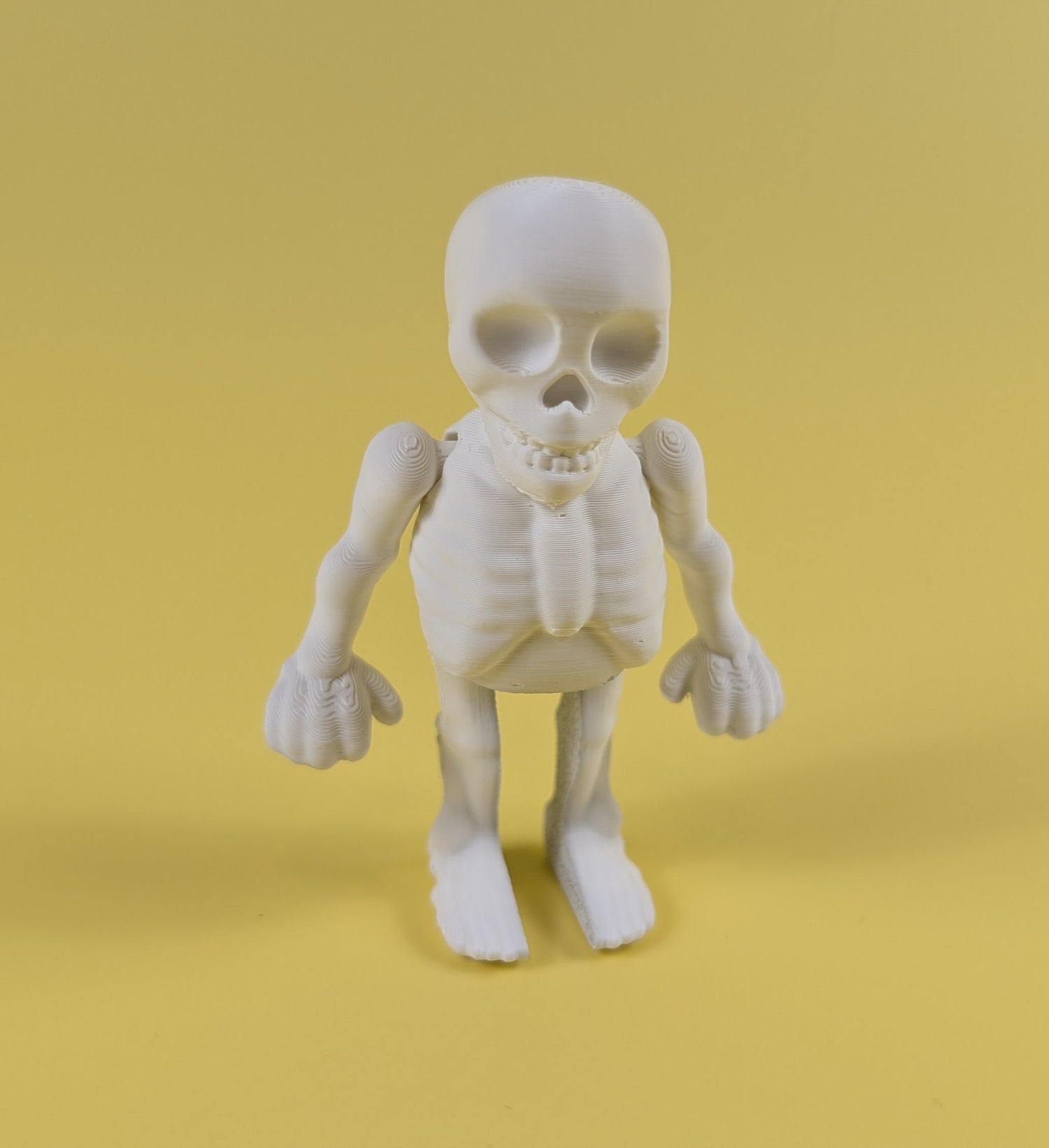 White 3D-printed skeleton figurine raising both arms on a yellow background; spooky-cute desk buddy with lightly poseable joints.