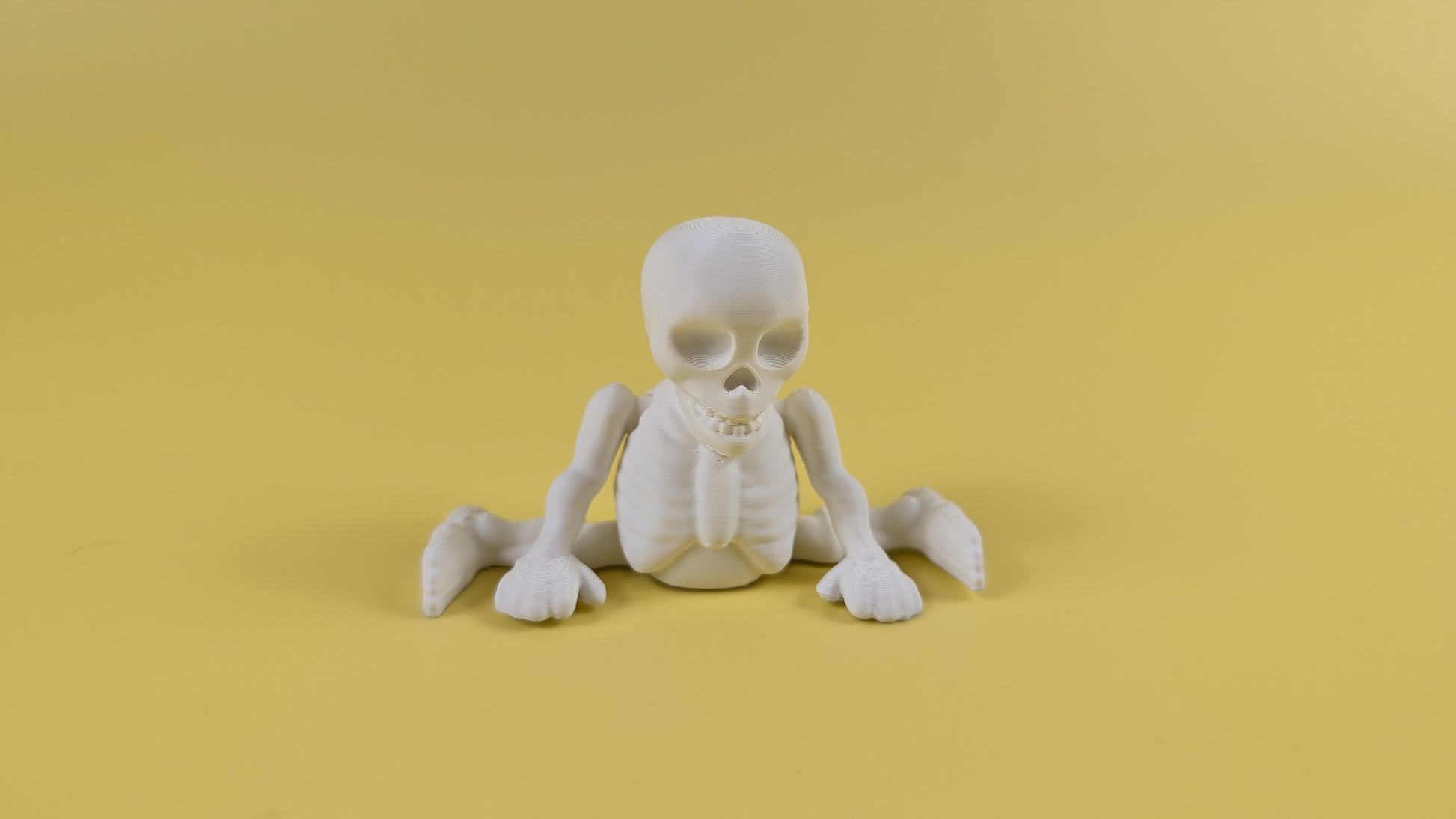 White 3D-printed skeleton figurine raising both arms on a yellow background; spooky-cute desk buddy with lightly poseable joints.
