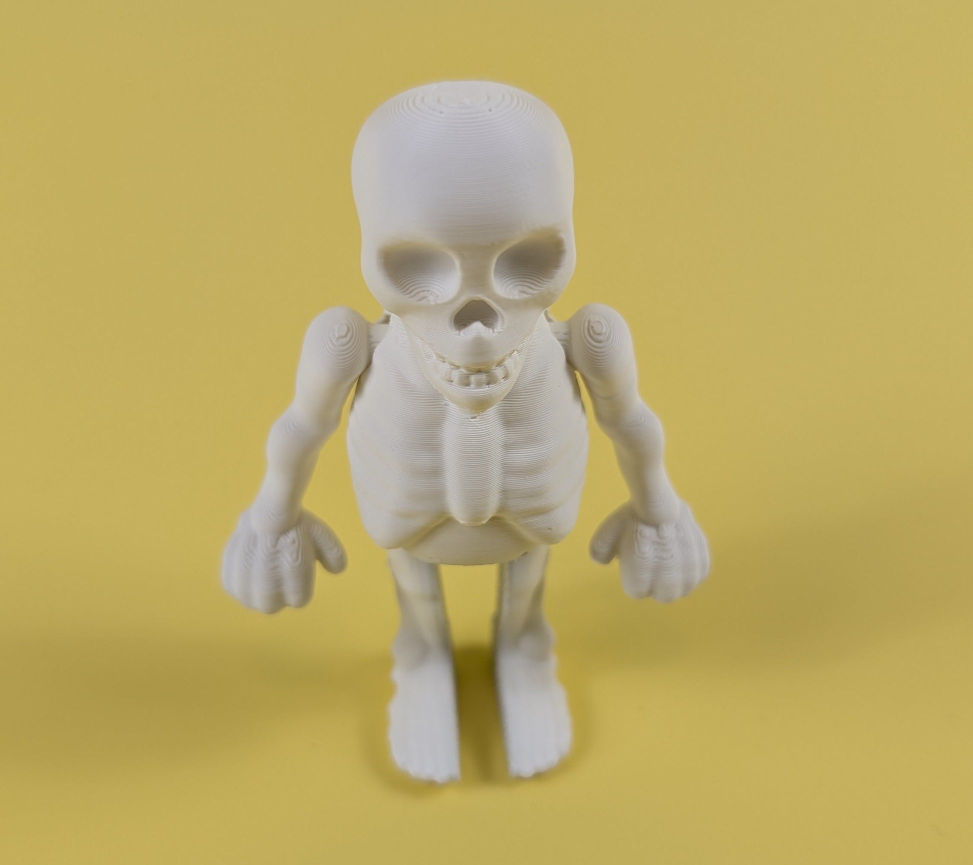 White 3D-printed skeleton figurine raising both arms on a yellow background; spooky-cute desk buddy with lightly poseable joints.
