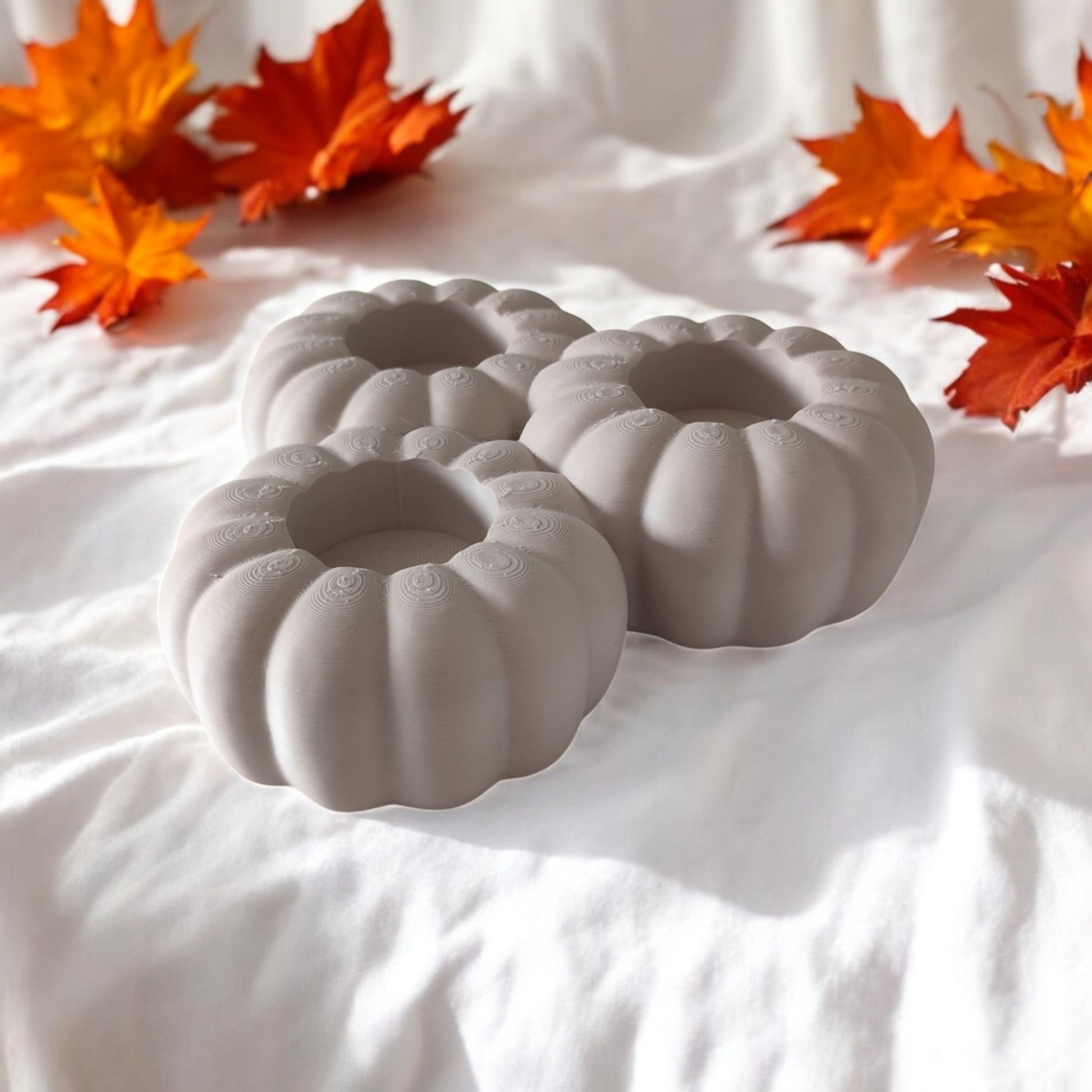 Set of three pumpkin tealight holders (16×14×5.5 cm), neutral colours, showing option with LED tealights lit, displayed on fall tabletop with autumn leaves.