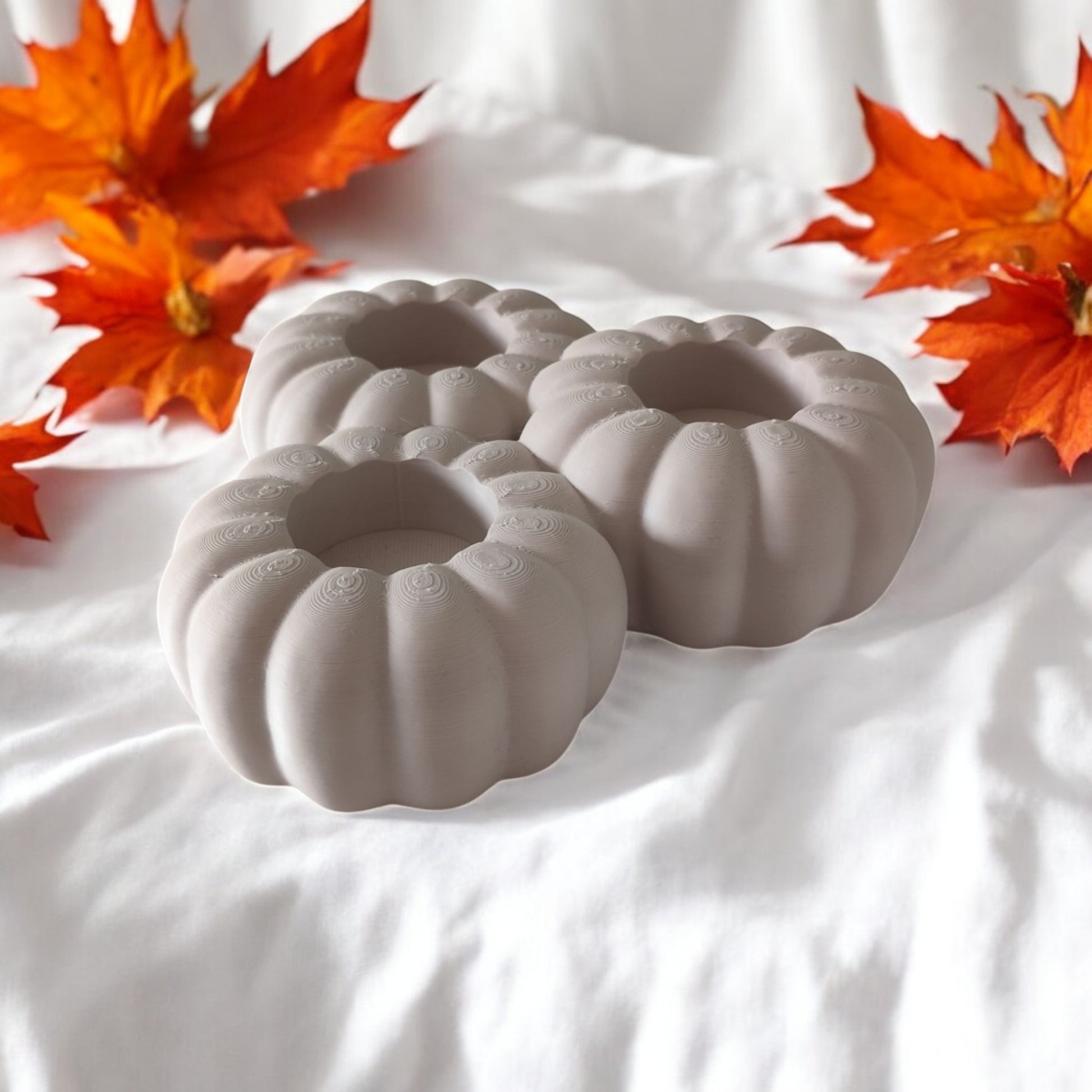 Set of three pumpkin tealight holders (16×14×5.5 cm), neutral colours, showing option with LED tealights lit, displayed on fall tabletop with autumn leaves.