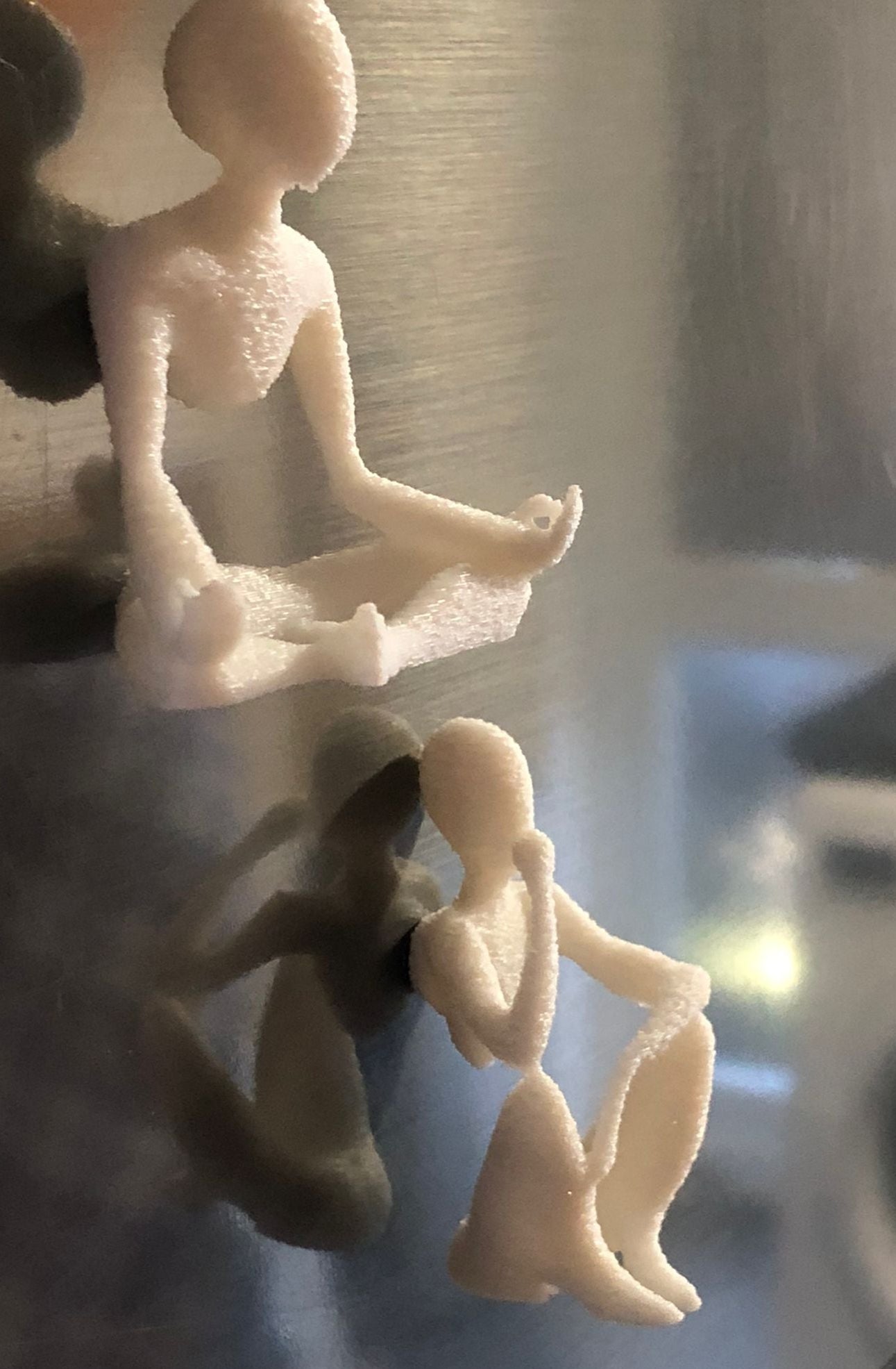 Set of two white 3D printed fridge magnets shaped like minimalist faceless human figures in yoga and meditation poses, displayed on a metallic surface.