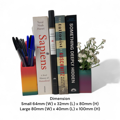 Colorful 3D printed rainbow-shaped organizer used as a bookend, pen holder, and mini vase, displayed in two sizes on a shelf with books, stationery, and dried flowers.