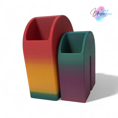 Colorful 3D printed rainbow-shaped organizer used as a bookend, pen holder, and mini vase, displayed in two sizes on a shelf with books, stationery, and dried flowers.