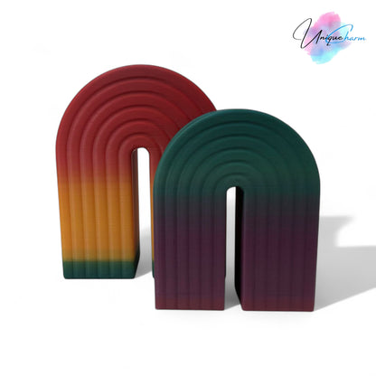Colorful 3D printed rainbow-shaped organizer used as a bookend, pen holder, and mini vase, displayed in two sizes on a shelf with books, stationery, and dried flowers.