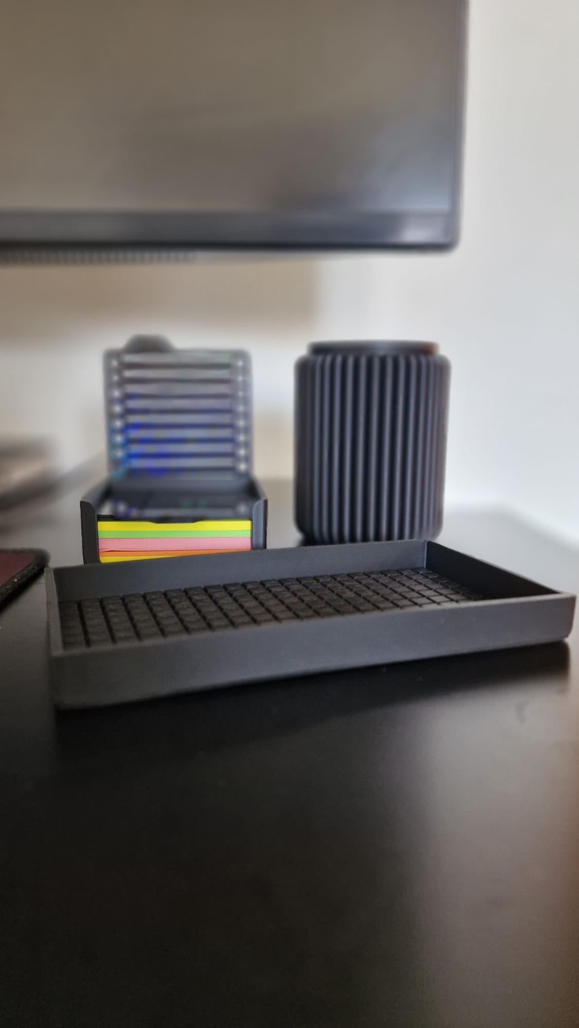 Modern black 3-piece desk organizer set including a ribbed pen holder, textured tray with earbuds case, and sticky notes holder with stencil cards on a black desk.
