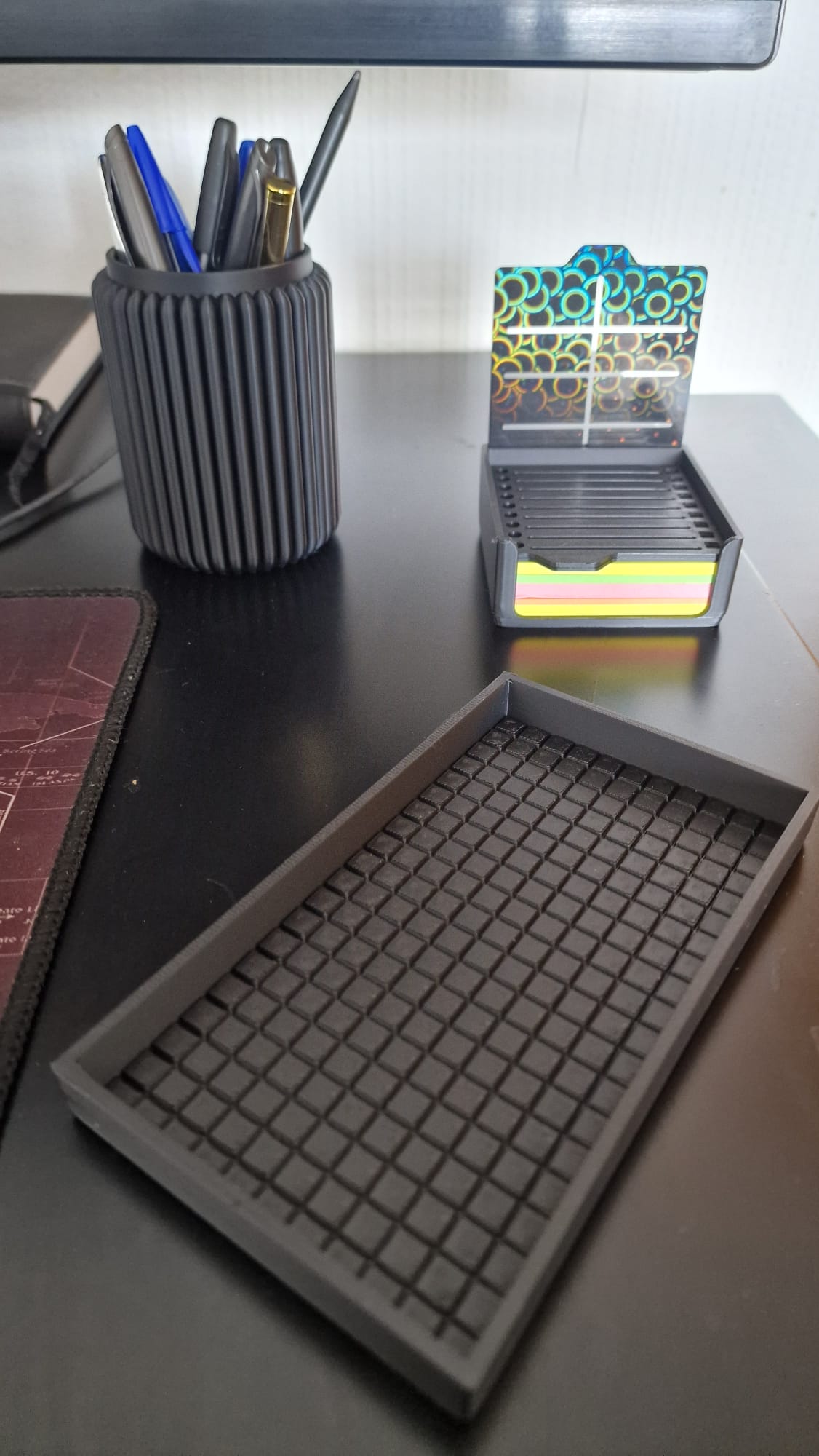 Modern black 3-piece desk organizer set including a ribbed pen holder, textured tray with earbuds case, and sticky notes holder with stencil cards on a black desk.