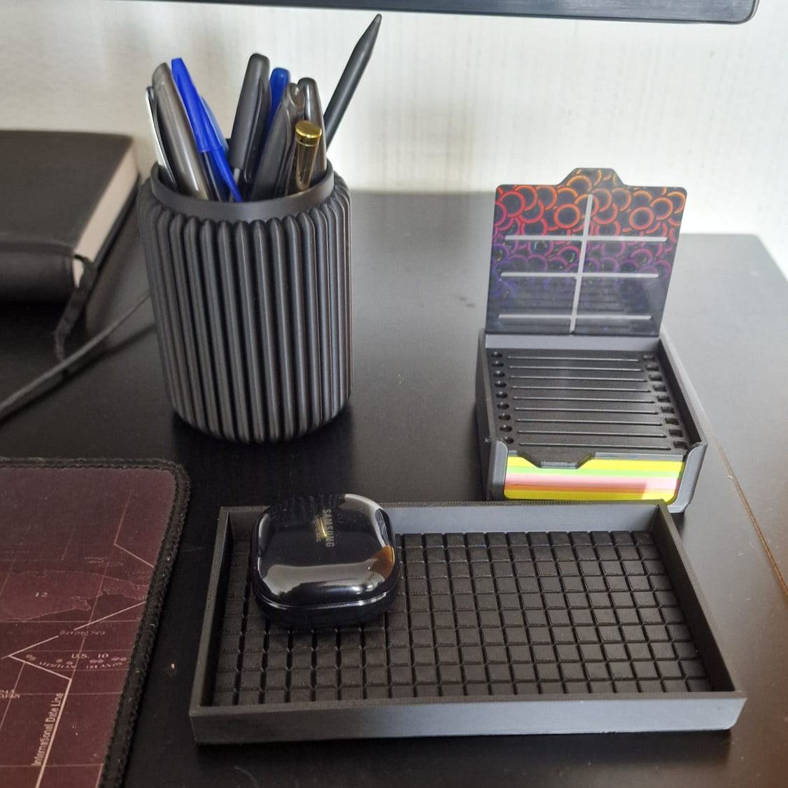 Modern black 3-piece desk organizer set including a ribbed pen holder, textured tray with earbuds case, and sticky notes holder with stencil cards on a black desk.