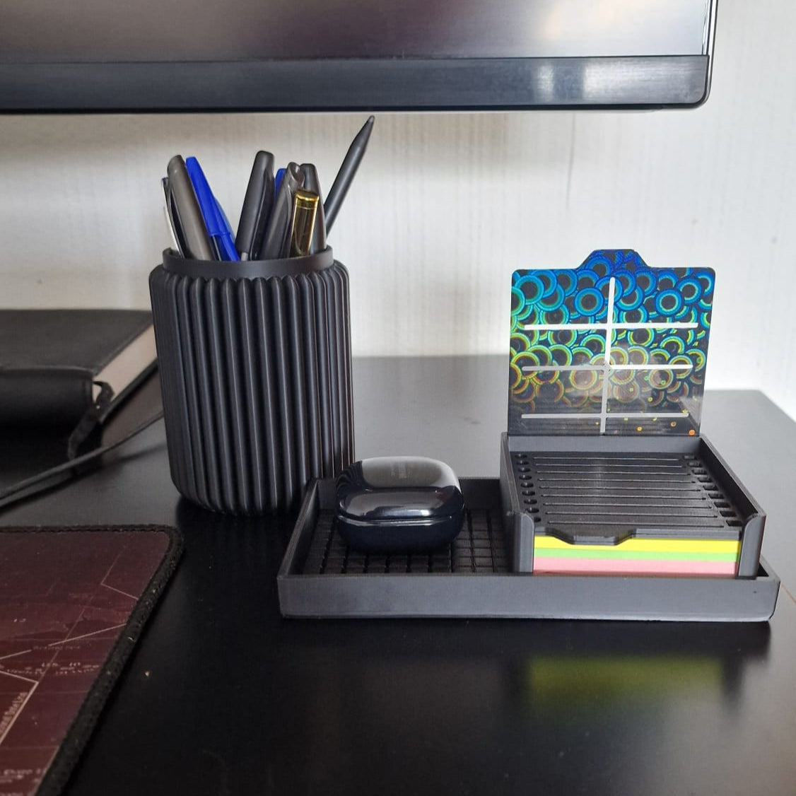 Modern black 3-piece desk organizer set including a ribbed pen holder, textured tray with earbuds case, and sticky notes holder with stencil cards on a black desk.