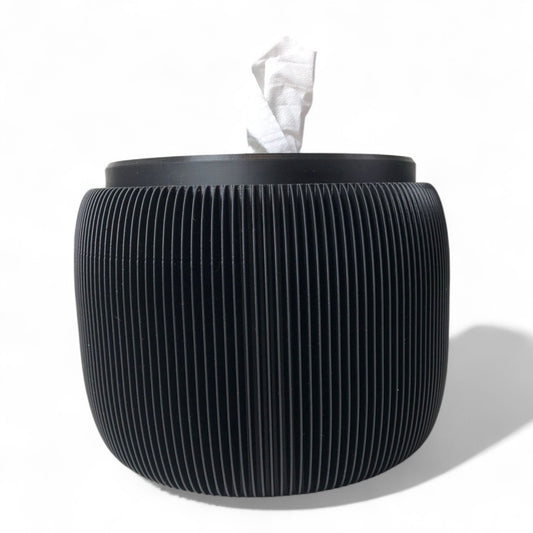 Modern black toilet paper holder with ribbed texture and lid, designed to prevent pets or children from unrolling the paper. Stylish and compact bathroom accessory shown on a white surface.