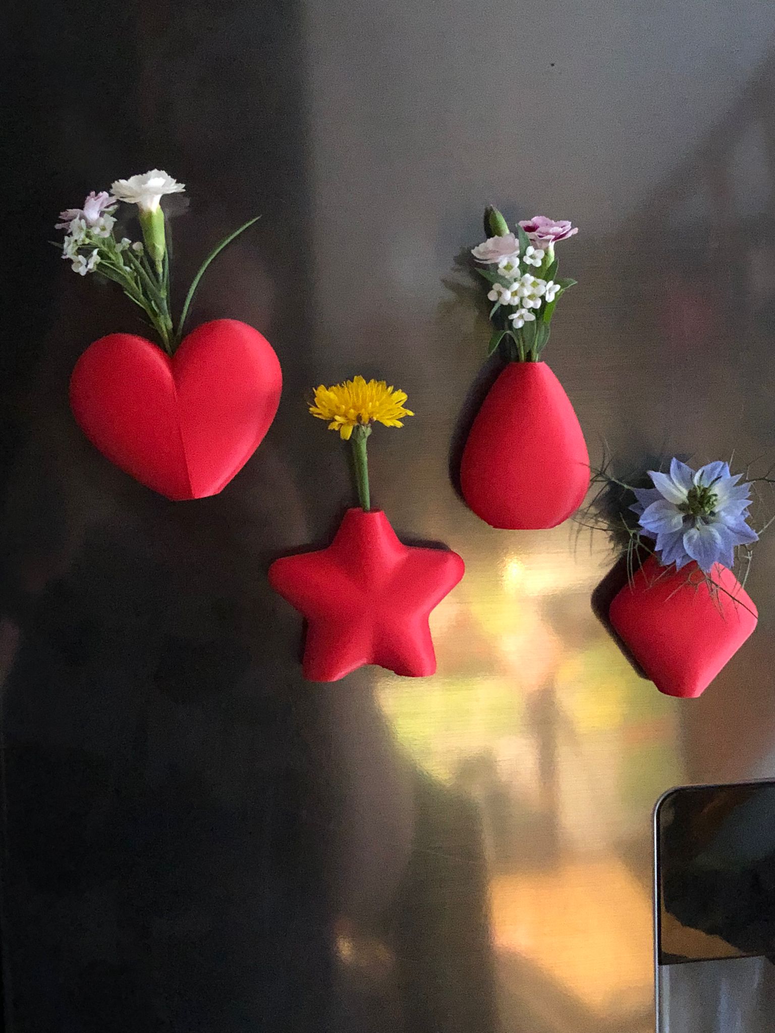Set of mini magnetic vases in heart, star, diamond, and teardrop shapes, attached to a refrigerator and holding small colorful flowers, displayed in red and white colors.