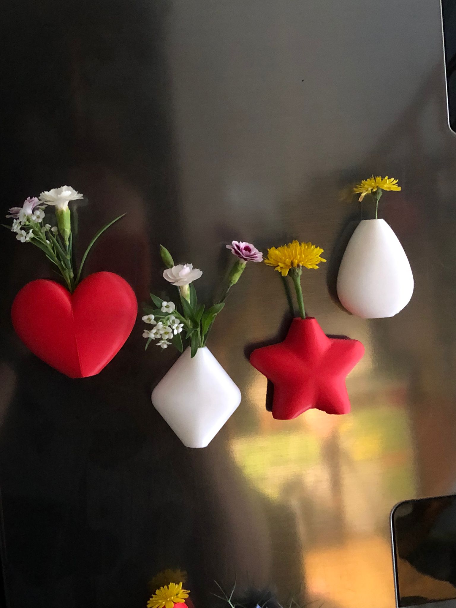 Set of mini magnetic vases in heart, star, diamond, and teardrop shapes, attached to a refrigerator and holding small colorful flowers, displayed in red and white colors.