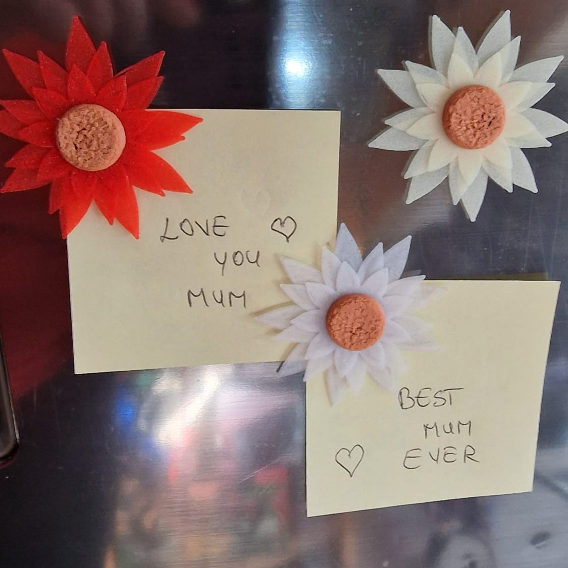 Two small notes with floral attachments on a reflective surface, one saying 'Love you Mum' and the other 'Best Mum Ever'.