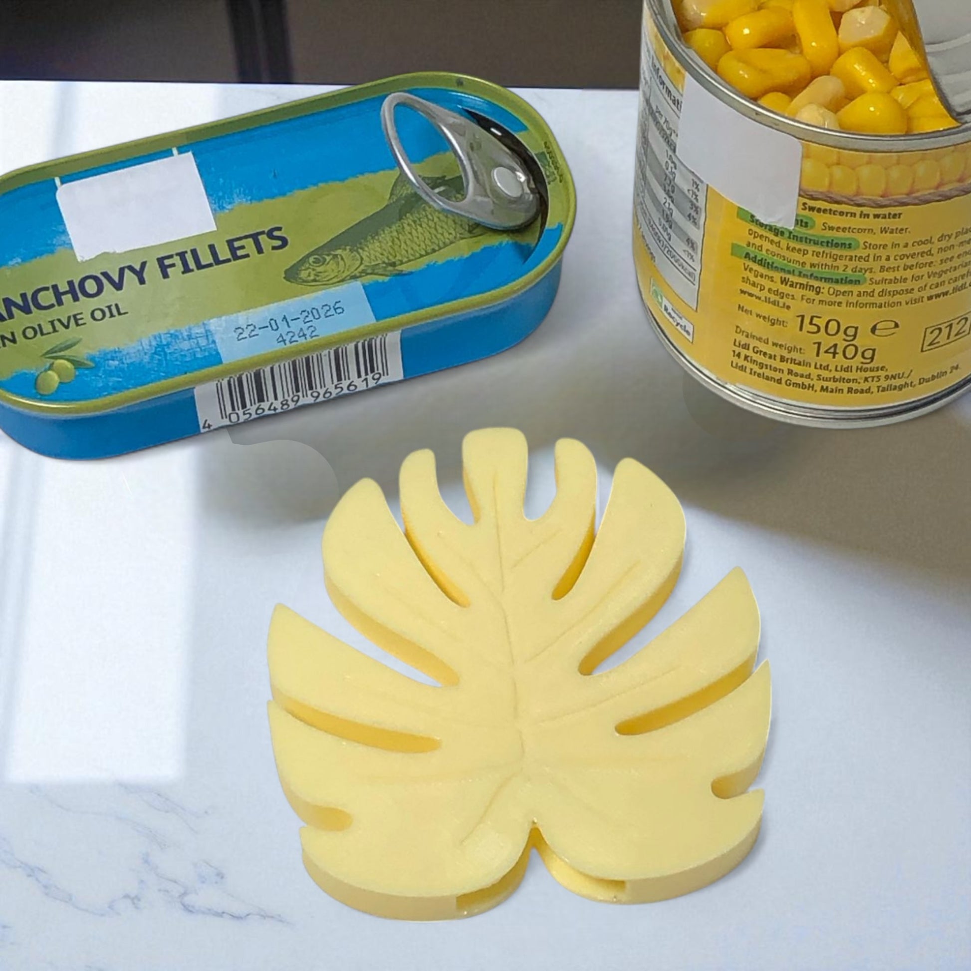 Canned food items including a can of anchovy fillets and a can of corn, with a leaf-shaped yellow object on a white surface.