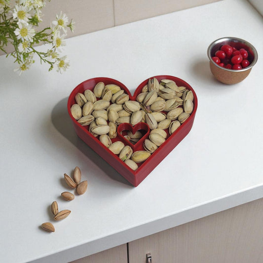 Heart-shaped red bowl filled with pistachios on a white surface with flowers and a small bowl of berries.