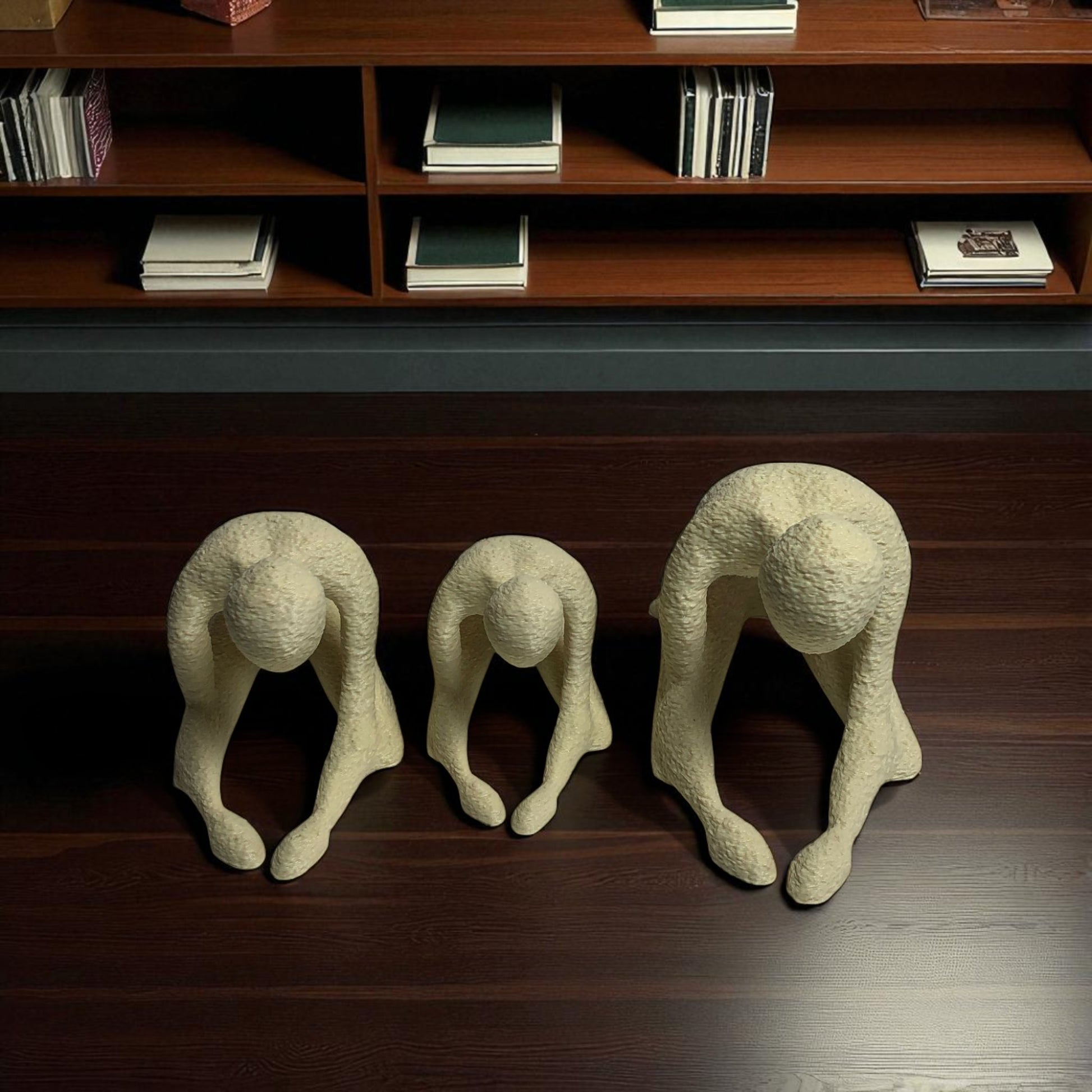 Three abstract stone sculptures of people in various poses on a wooden floor with a bookshelf in the background.