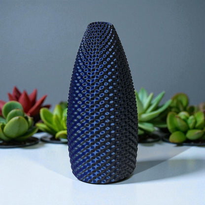 Textured blue and black vase on a white surface with plants in the background