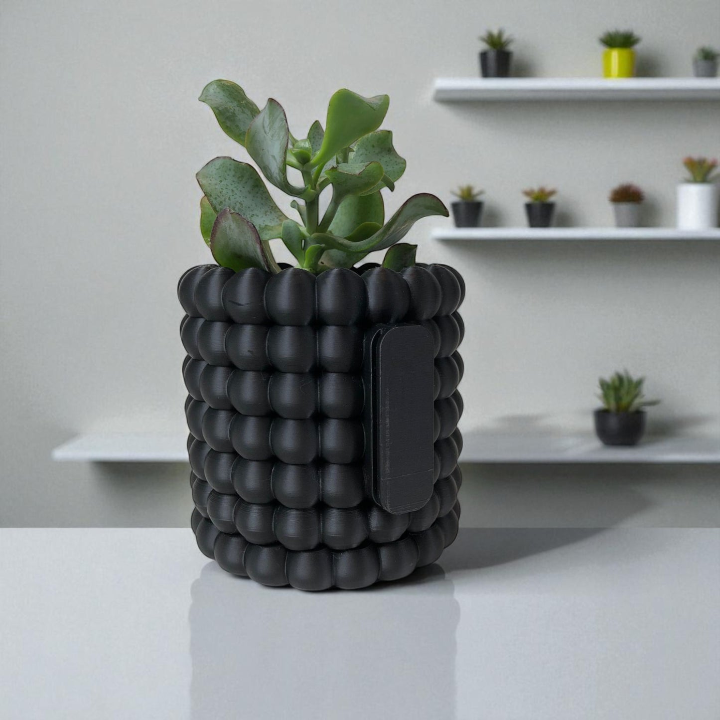 modern black wall-mounted planter with a bubble texture design, holding a green succulent plant. includes mounting bracket for screws or adhesive and a built-in water tray.