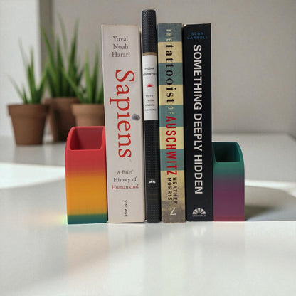Colorful 3D printed rainbow-shaped organizer used as a bookend, pen holder, and mini vase, displayed in two sizes on a shelf with books, stationery, and dried flowers.