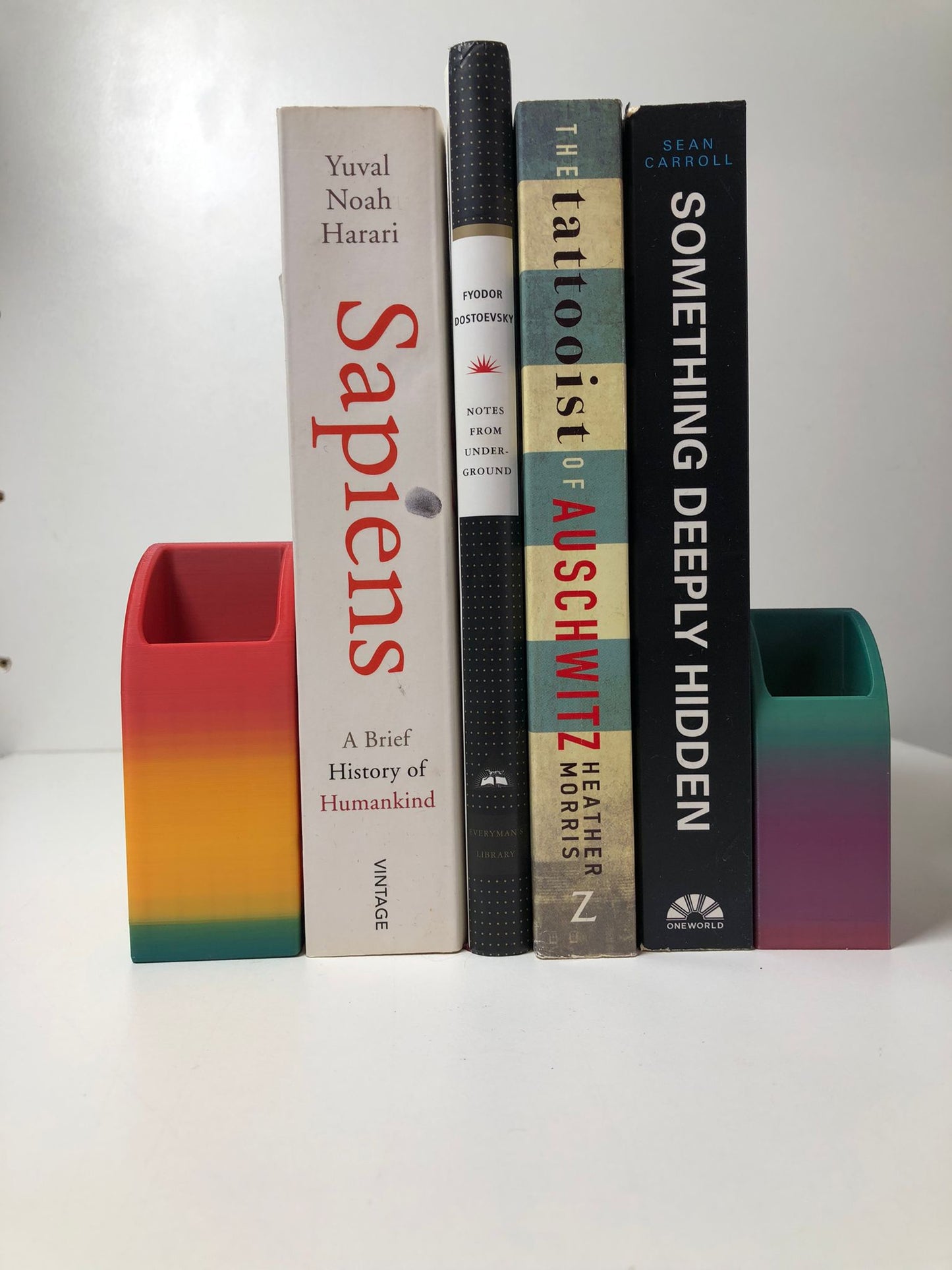 colorful 3d printed rainbow-shaped organizer used as a bookend, pen holder, and mini vase, displayed in two sizes on a shelf with books, stationery, and dried flowers.