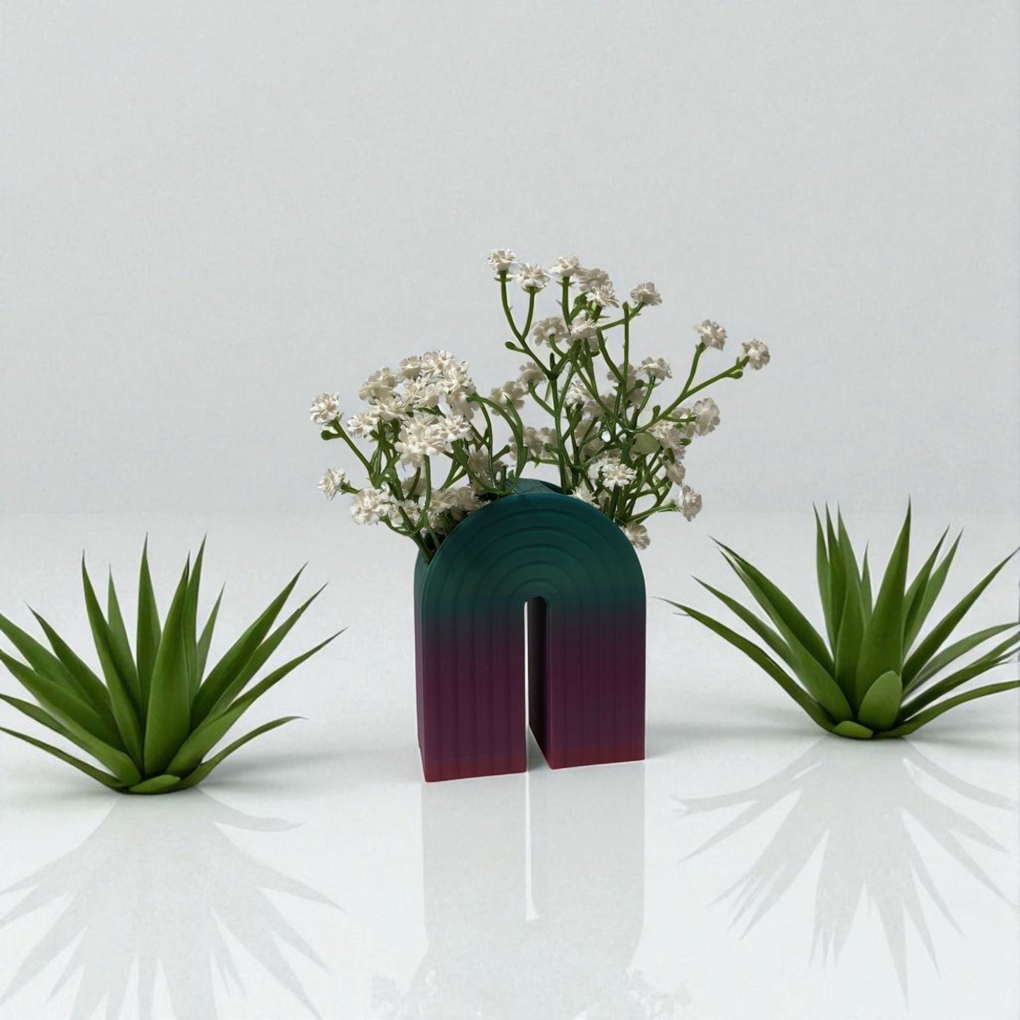 colorful 3d printed rainbow-shaped organizer used as a bookend, pen holder, and mini vase, displayed in two sizes on a shelf with books, stationery, and dried flowers.