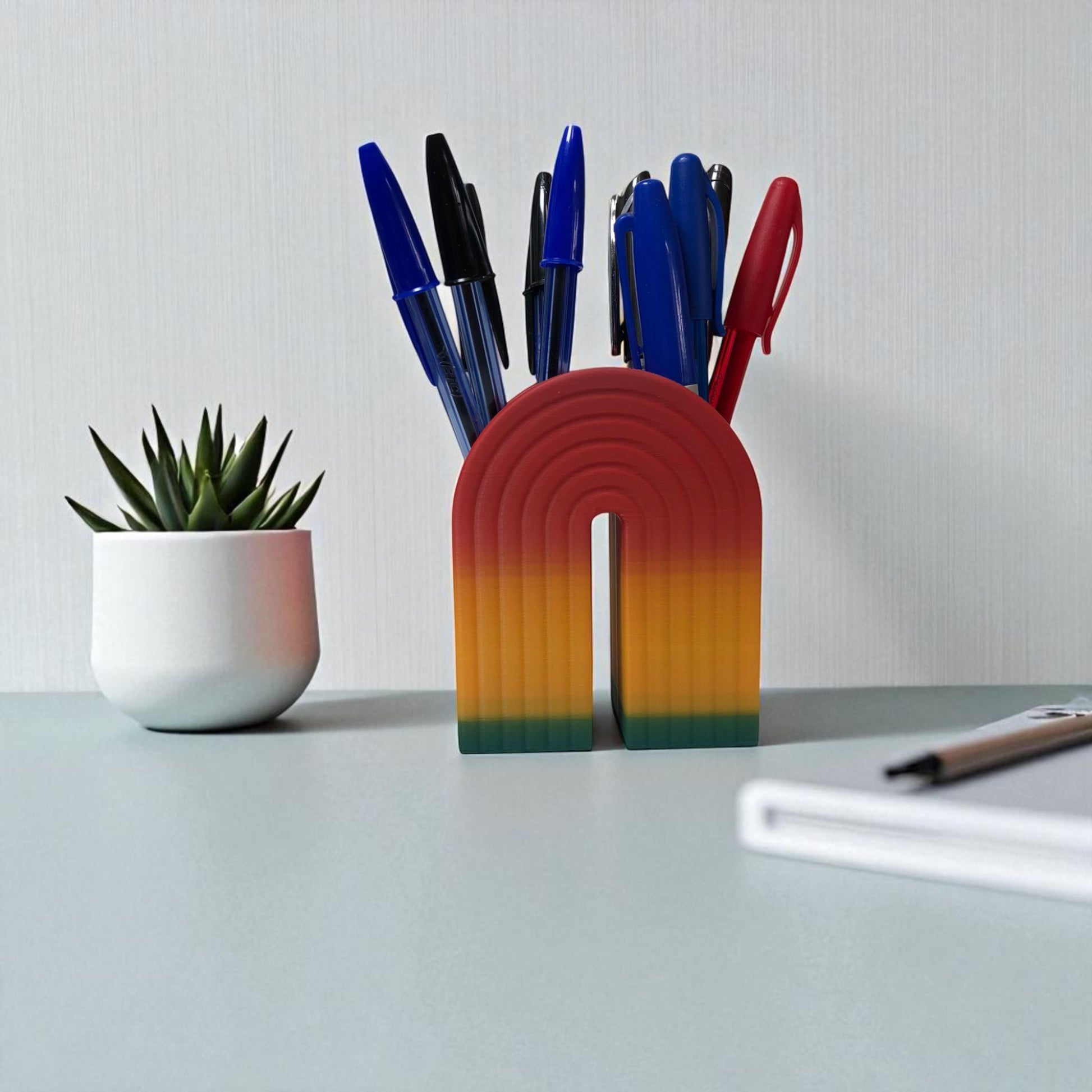 Colorful 3D printed rainbow-shaped organizer used as a bookend, pen holder, and mini vase, displayed in two sizes on a shelf with books, stationery, and dried flowers.