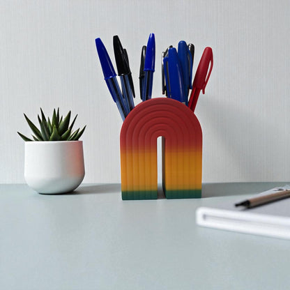 Colorful 3D printed rainbow-shaped organizer used as a bookend, pen holder, and mini vase, displayed in two sizes on a shelf with books, stationery, and dried flowers.