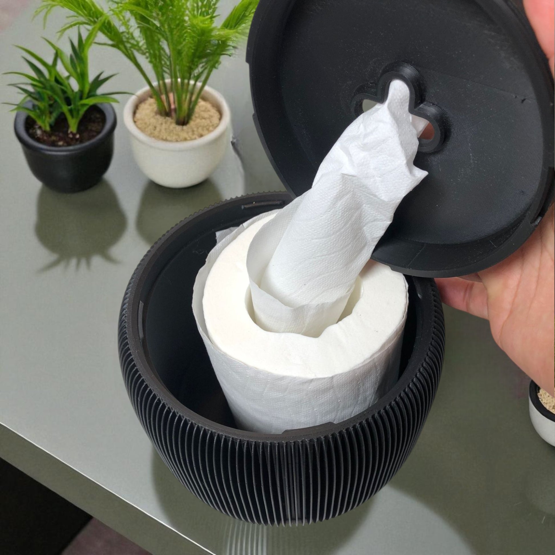 Modern black toilet paper holder with ribbed texture and lid, designed to prevent pets or children from unrolling the paper. Stylish and compact bathroom accessory shown on a white surface.
